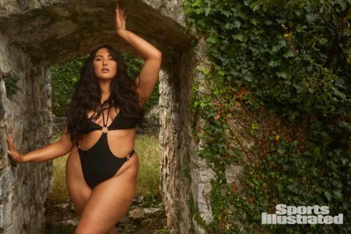 Yumi Nu stands under an ivy-covered stone archway in a black one-piece with front cut-outs.